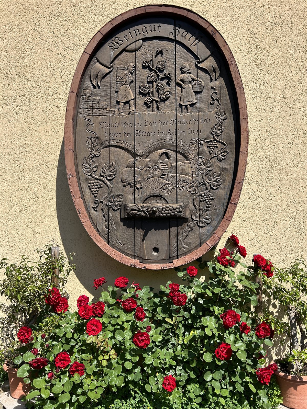 Geschnitztes ovales Holzrelief mit der Aufschrift Weingut Zaiß, Weinbauszenen und Trauben, darunter rote Geranien